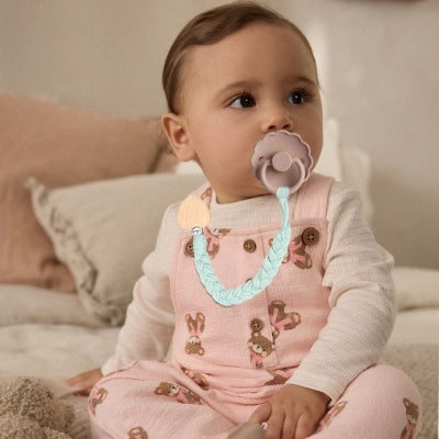 Attache-tétine-turquoise-coeur-bois bébé assis sur un lit habillé en rose motif ours brun 