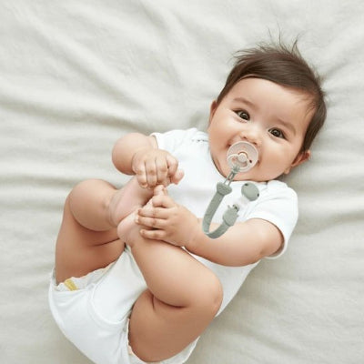 Attache tetine motif girafe vert bébé souriant couché sur son lit joue ave cses pieds