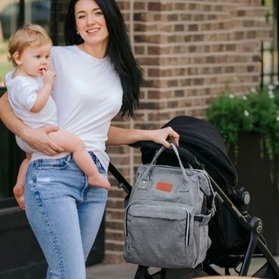 Sac-à-dos-à-langer-gris-poussette-bebe-dans-les-bras-de-maman-souriante-mur-brique